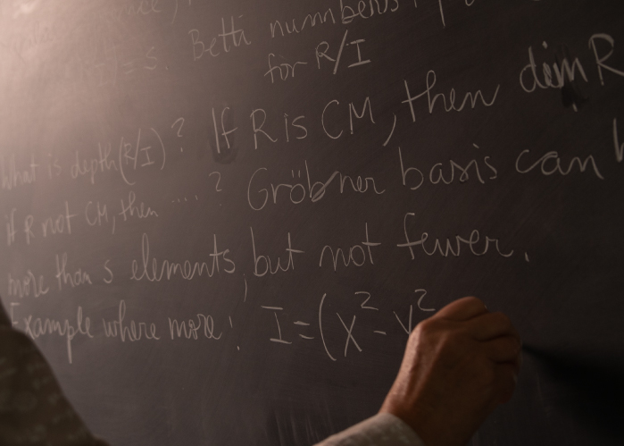 Close-up photo of a person’s hand writing math equations on a blackboard