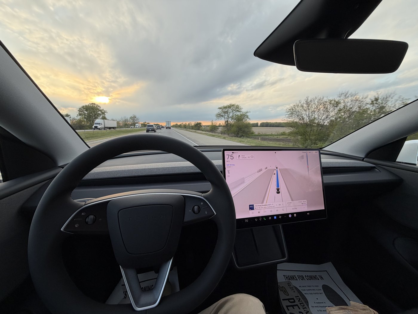 View from the driver's seat of a Tesla on a two-lane highway at sunset. Nobody's hands are on the steering wheel in the foreground. The center touchscreen shows Self-Driving engaged at 75 mph, with the car rendered centered in its lane and surrounding traffic displayed ahead.